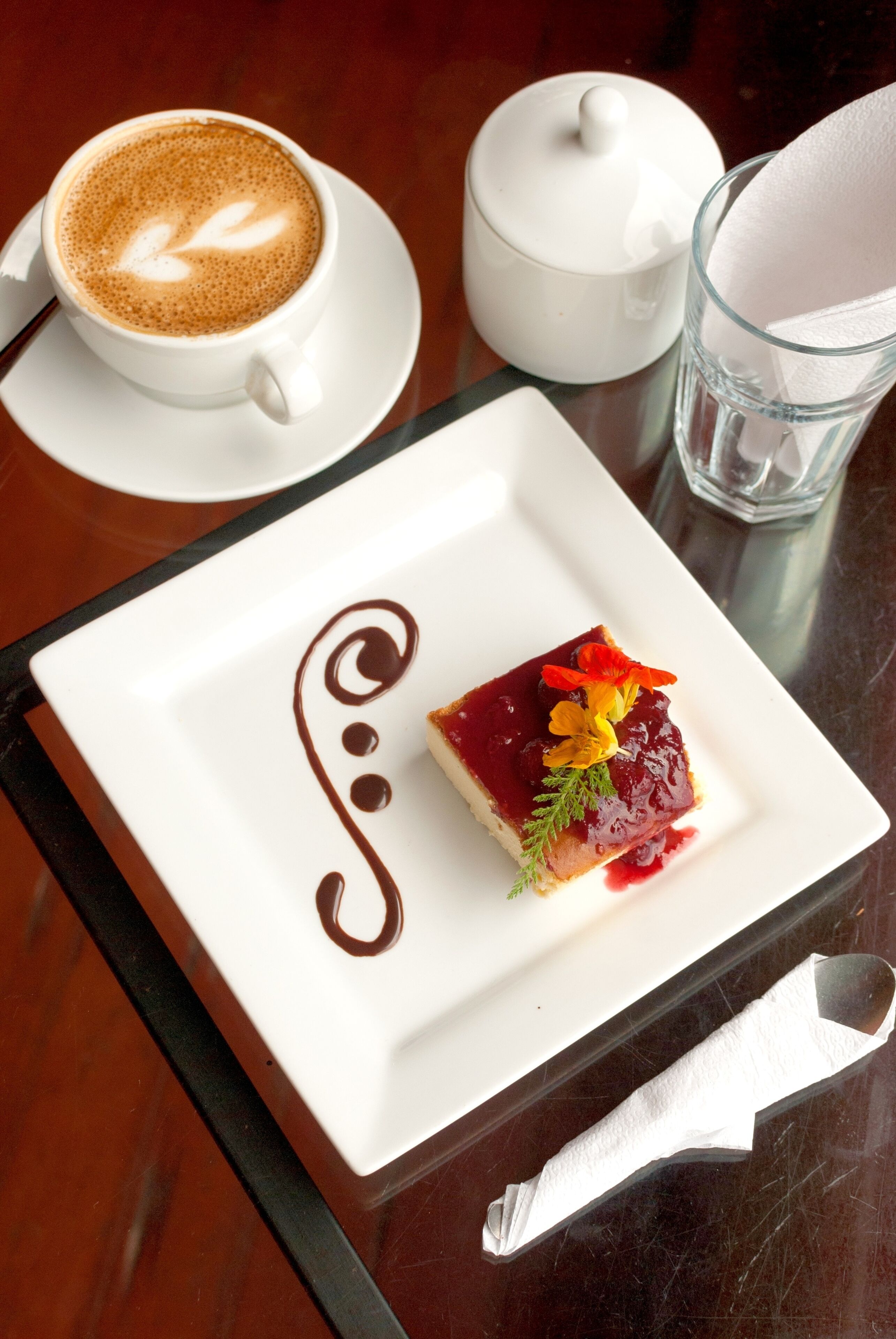 Un postre sobre un plato blanco con un diseño de chocolate decorativo, una taza de café, un azucarero y un vaso de agua sobre una mesa de madera.
