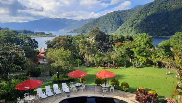 Un área de piscina con sillones de descanso y sombrillas rojas, rodeada de abundante vegetación y vistas a un lago y montañas.