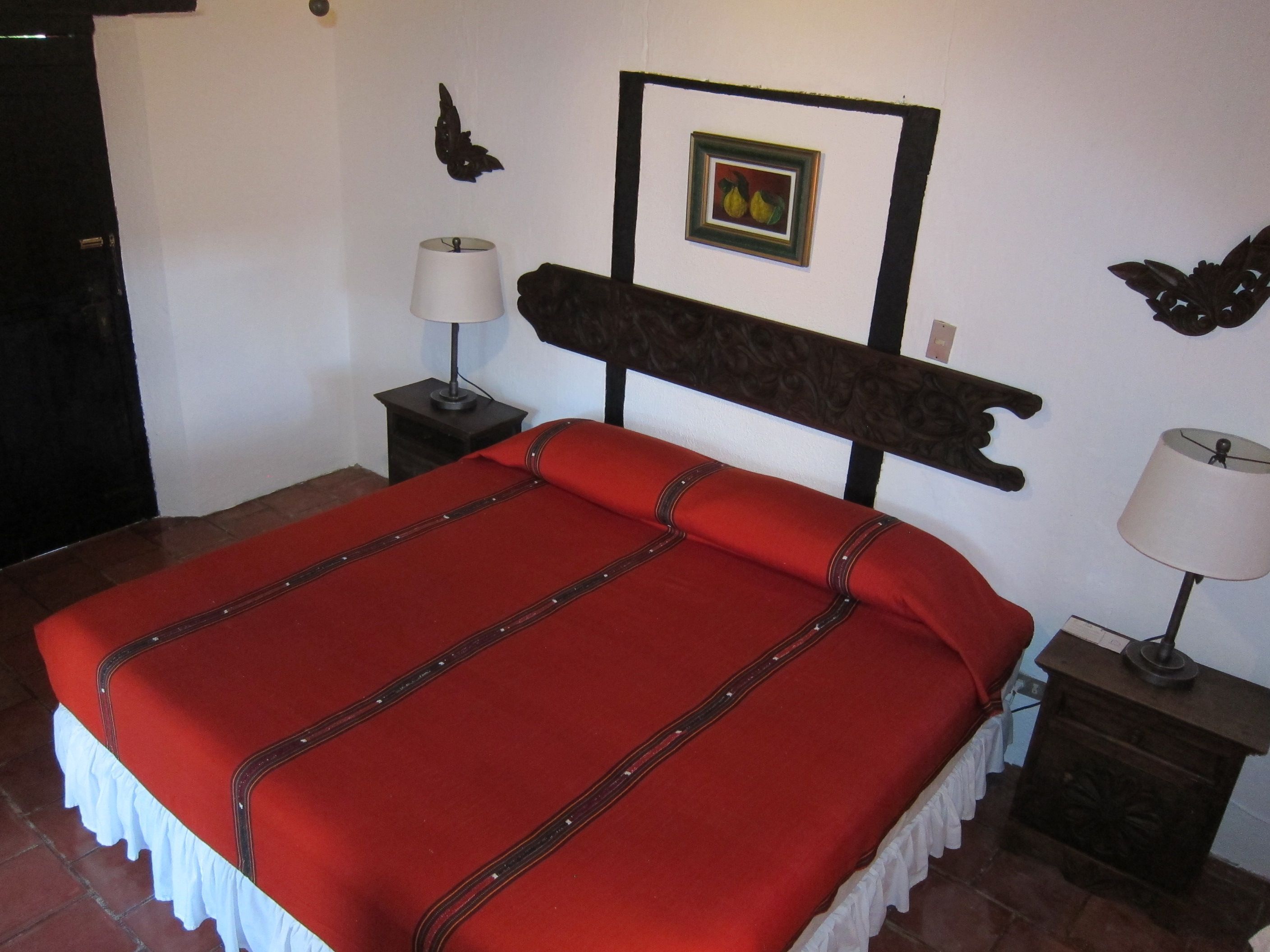 Ein Bett mit roter Bettdecke und weißer Bordüre, zwei Nachttischlampen, ein gerahmtes Bild an der Wand und ein dekoratives Wandbild.