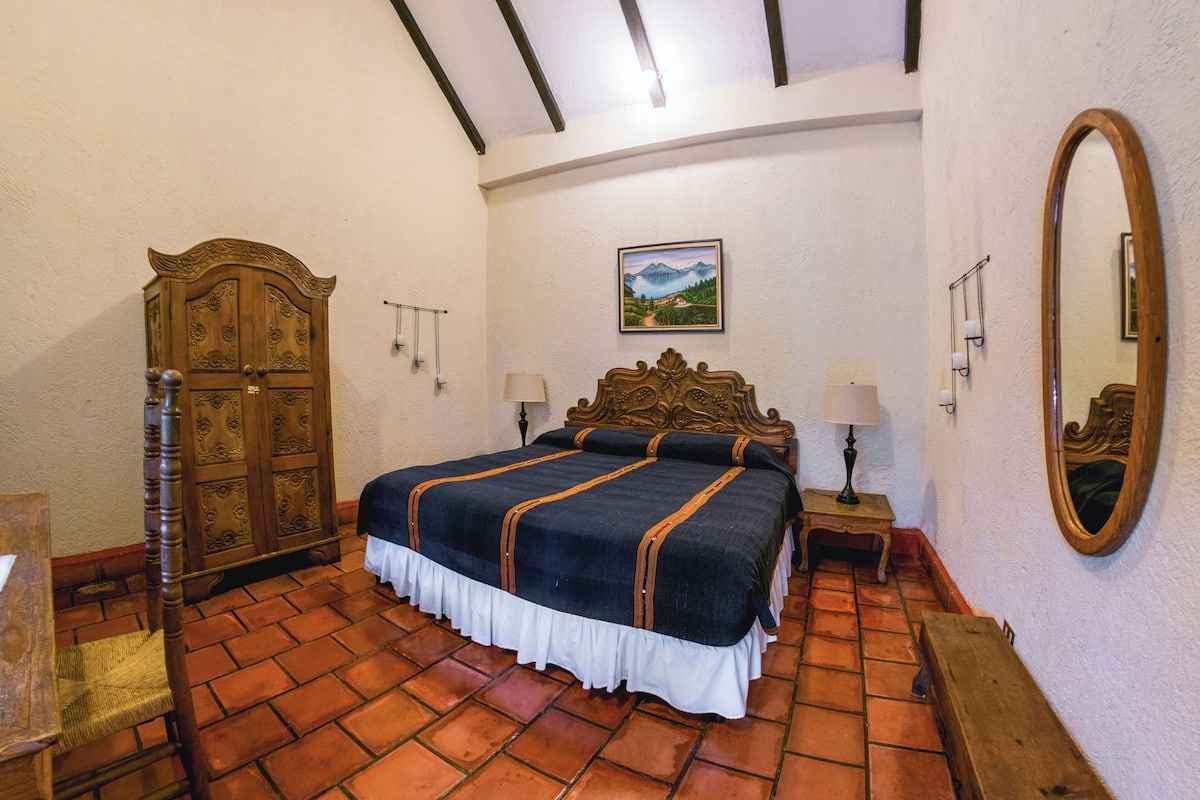 Un dormitorio con una cama de madera, un armario de madera, una mesita de noche con una lámpara, un espejo y un cuadro en la pared.