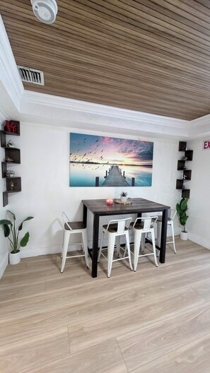 A dining area with a wooden ceiling, a large painting of a sunset over a pier, and a white table with chairs.