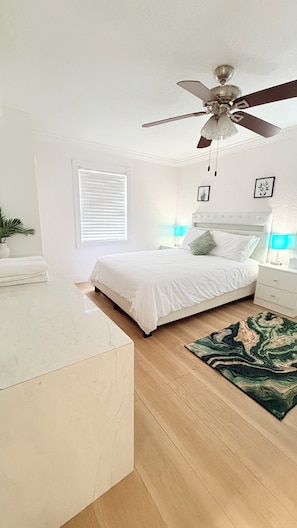 Un dormitorio con cama, ventilador de techo, mesita de noche con lámpara, ventana con persianas y una planta.
