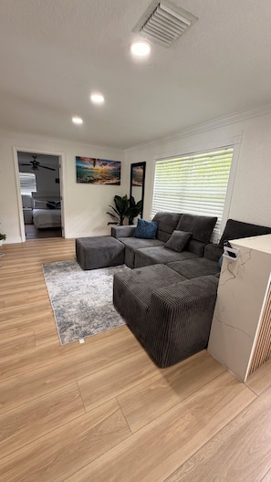 A modern living room with a sectional sofa, a grey rug, wooden flooring, a ceiling fan, and a painting on the wall.