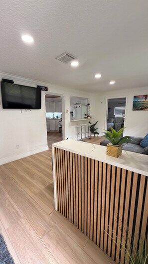 A modern living room with a wooden slatted partition, a flat-screen TV mounted on the wall, and a kitchen area in the background. - Entire House Climatized Pool Close to the Beach (Hollywood)