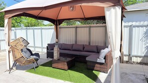 A covered outdoor seating area with a sofa and chairs, a small table, and artificial grass.
