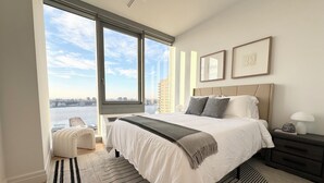 A modern bedroom with a large window, a bed with a grey throw, a bedside table with a lamp, and a view of the city. - 2 bed/2 bath luxe condo in Midtown  (New York)