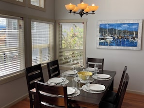 Un coin repas avec une table dressée pour quatre personnes, quatre chaises, un lustre et un tableau représentant un paysage portuaire.