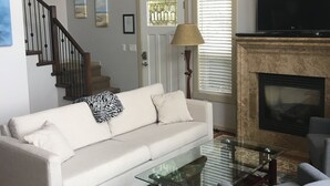 A living room with a white sofa, a glass coffee table, a fireplace, a television, and a staircase.