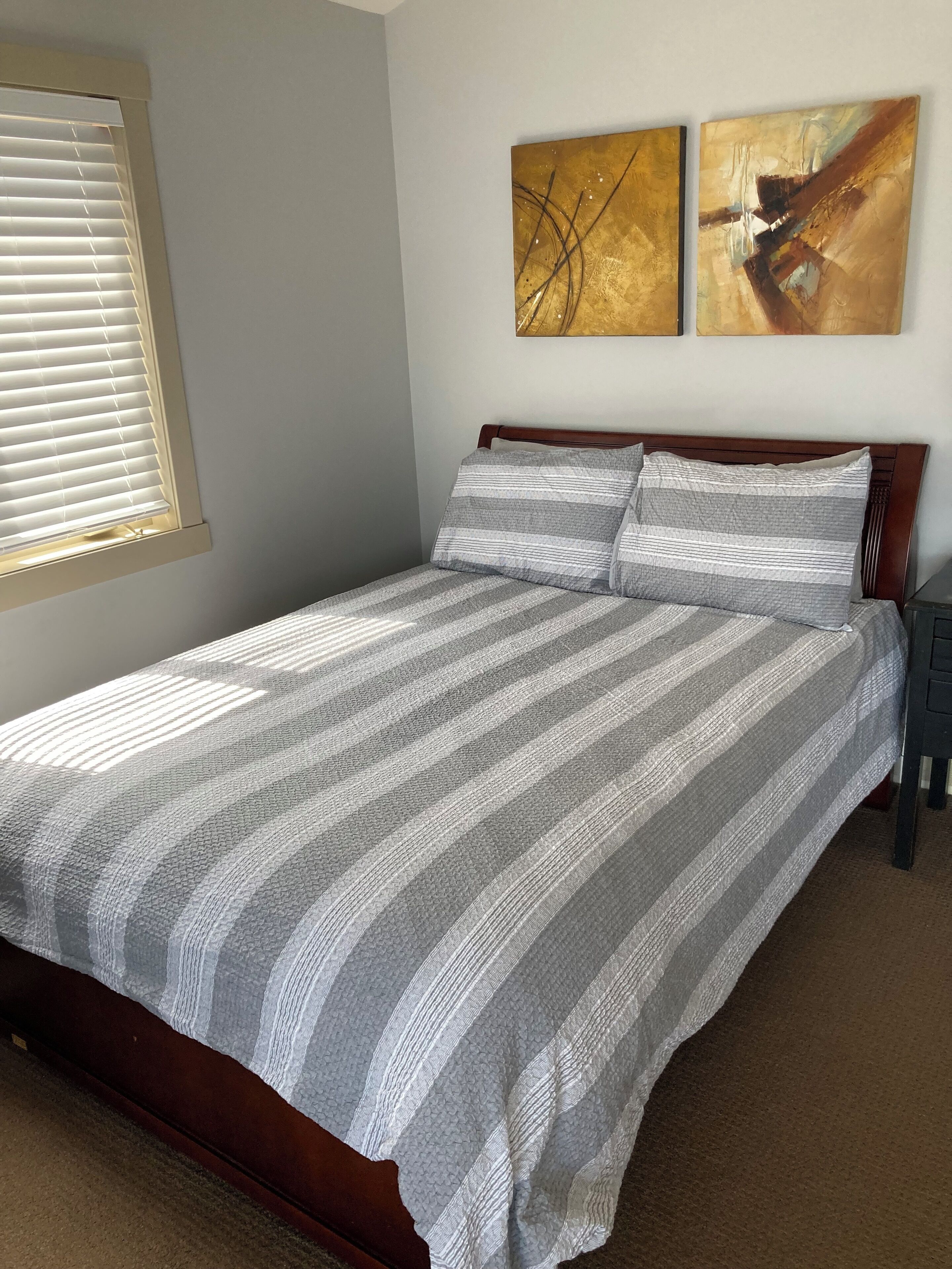 A neatly made bed with a striped bedspread and two pillows, a window with blinds, and two abstract paintings on the wall.