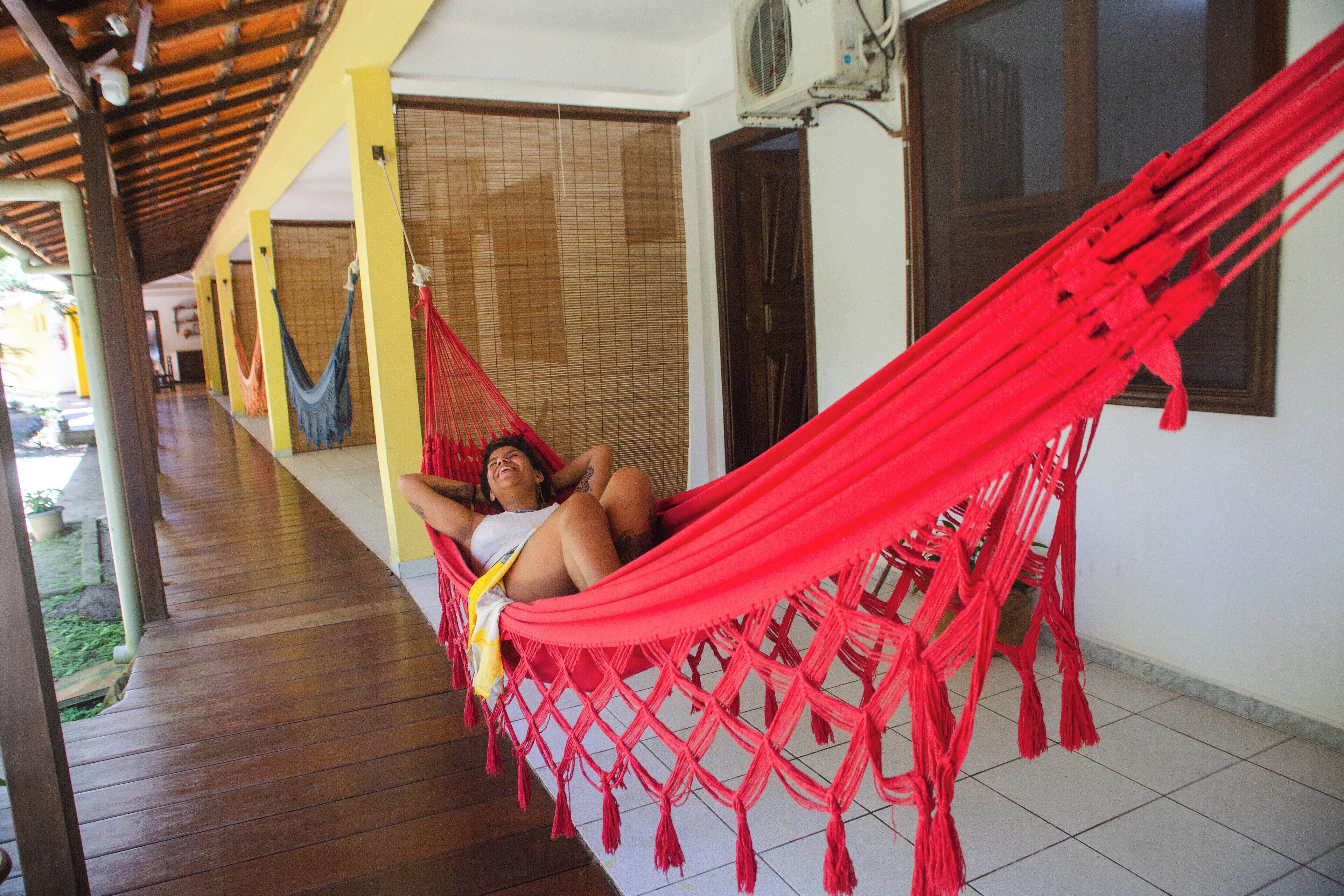 Una persona descansando en una hamaca roja en un porche de madera con piso de baldosas, una pared amarilla y una ventana con persianas.