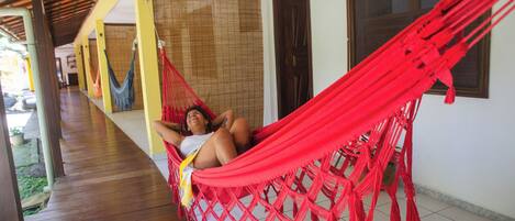 A person relaxing in a red hammock on a wooden porch with a tiled floor, a yellow wall, and a window with blinds.