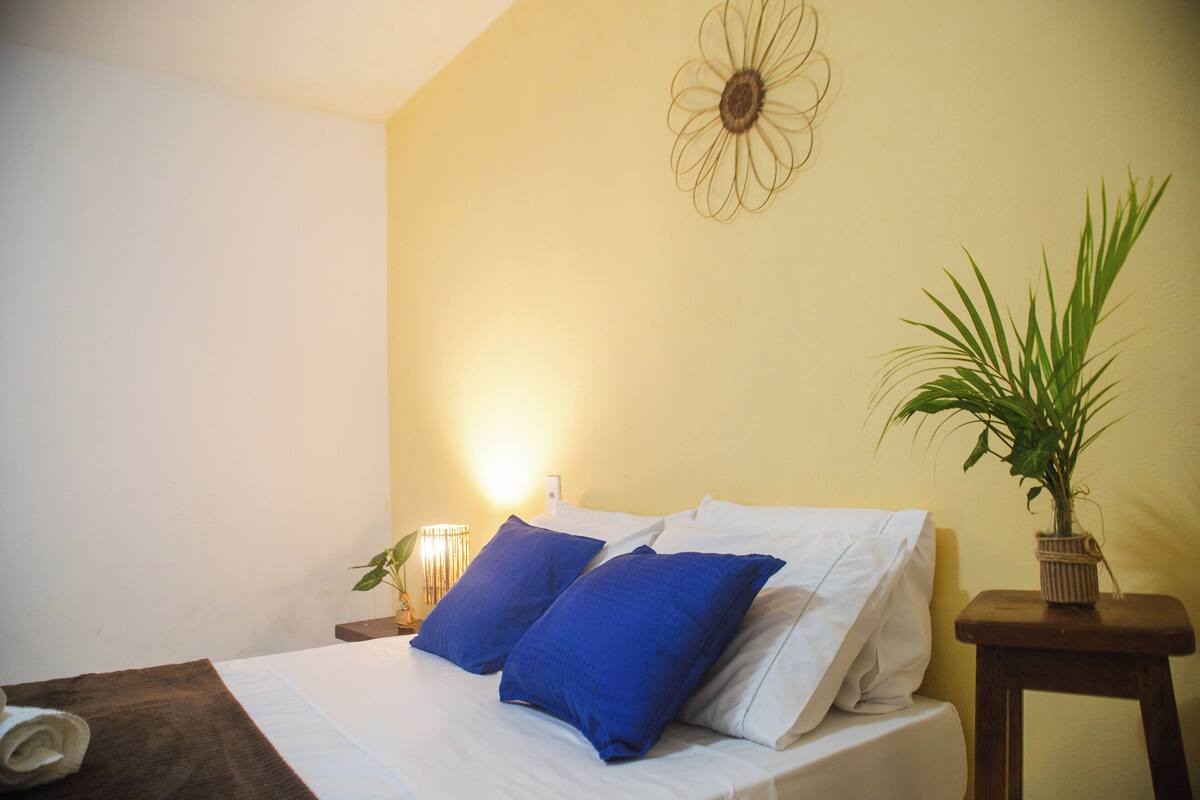 A neatly made bed with white and blue pillows, a small bedside table with a lamp, and a potted plant on a wooden nightstand.