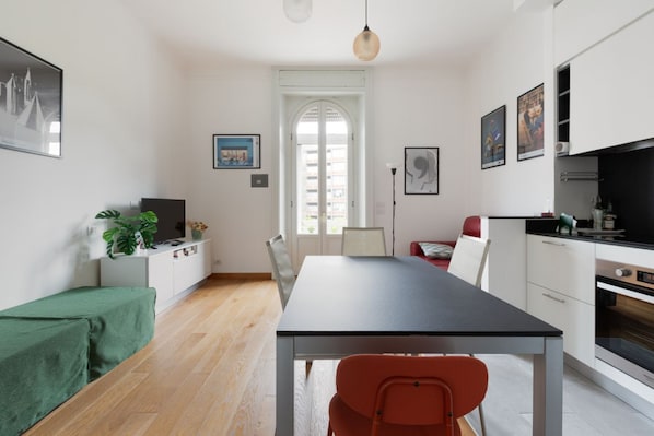 A modern kitchen with white cabinets, a dining table, and chairs. There is a window with a view of buildings outside.
