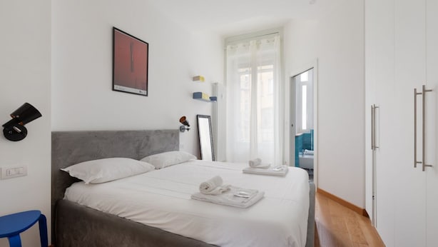 A bedroom with a bed, a chair, a bedside table, a mirror, and a window with curtains. - GuestHost - Buschi Milano-Città Studi Apartment (Milano)