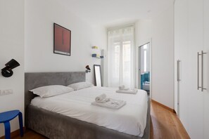 Ein Schlafzimmer mit einem Bett, einem Sessel, einem Nachttisch, einem Spiegel und einem Fenster mit Vorhängen.