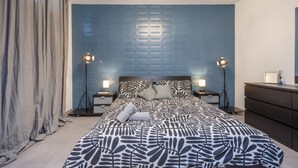 Une chambre à coucher avec une couvre-lit à motifs, une tête de lit à motifs géométriques, deux lampes de chevet et une commode sombre.