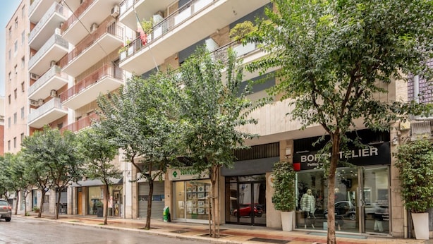Un immeuble à plusieurs étages, avec une boutique au rez-de-chaussée, des arbres bordant le trottoir et une voiture garée.