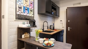 Une cuisine compacte avec un plan de travail foncé, un évier et une machine à café. Un poster encadré est accroché au mur, et une porte se trouve sur la droite.
