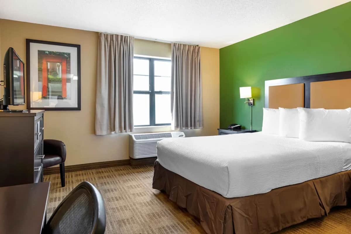 Une chambre d’hôtel avec un grand lit, un bureau, une chaise, un tableau encadré au mur et une fenêtre avec des rideaux.