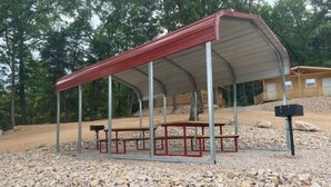 Un área de picnic techada con bancos y un refugio, rodeada de grava y árboles.