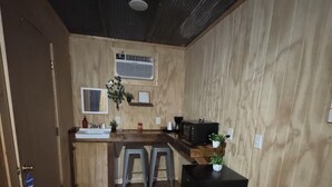 Una cocina pequeña con encimera de madera, un fregadero, un microondas y un cubo de basura. Hay dos taburetes y una planta en maceta.