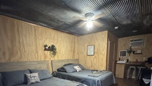 Una habitación con dos camas, una pared de madera, un ventilador de techo, una pequeña kitchenette y una puerta.
