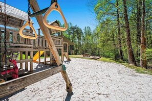 A wooden play structure with swings and a slide, surrounded by a forested area and a picnic table.