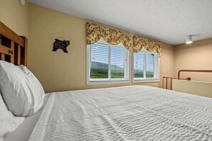 Une chambre à coucher avec un grand lit, deux fenêtres équipées de stores, une tête de lit en bois et un panneau décoratif fixé au mur.