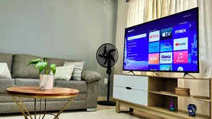 A living room with a grey sofa, a wooden coffee table with a plant, a standing fan, and a TV displaying Roku TV interface. - Oferta Excelente Casa En Residencial Privada, Alberca, Asador (Ciudad Valles)
