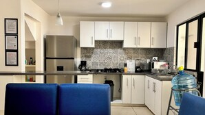 A modern kitchen with white cabinets, a stainless steel refrigerator, a coffee maker, and a water dispenser. There is a dining area with blue chairs and a table. - Oferta Excelente Casa En Residencial Privada, Alberca, Asador (Ciudad Valles)