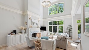 A spacious living room with a fireplace, comfortable seating, and large windows offering a view of greenery.