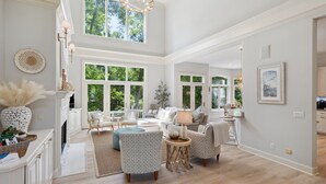 A spacious living room with a high ceiling, large windows, and a fireplace. There are multiple seating areas with sofas and chairs, and a dining table.