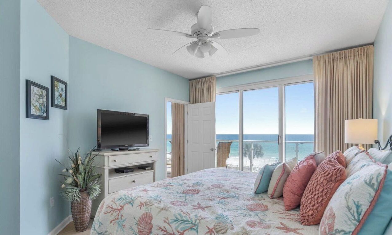 A bedroom with a bed, a television, a plant, and a view of the ocean.