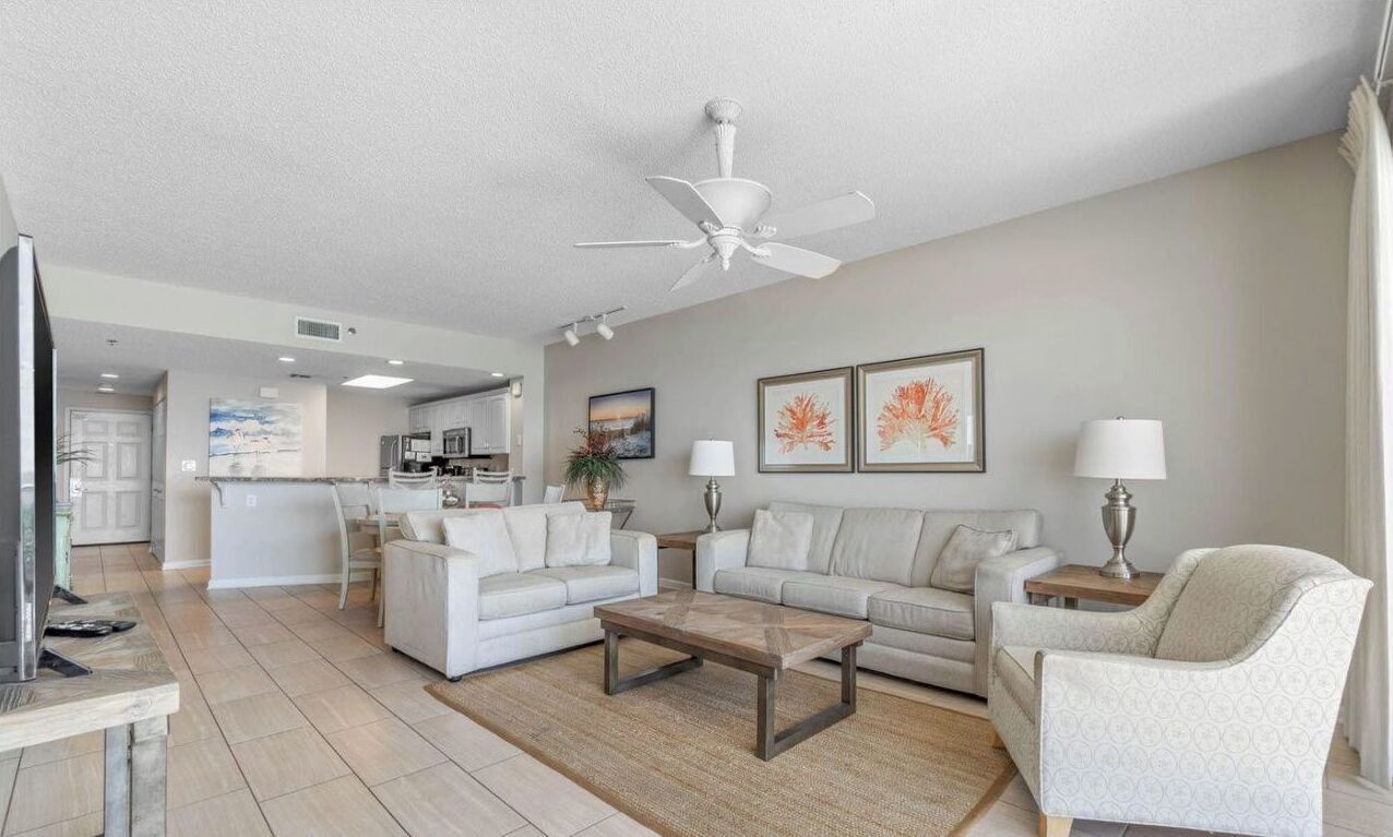 A spacious living room with a large white ceiling fan, two sofas, a coffee table, and a TV. A kitchen is visible in the background.
