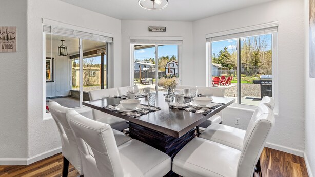 A modern dining area with a large rectangular table, white upholstered chairs, and a view of the outdoors through large windows.