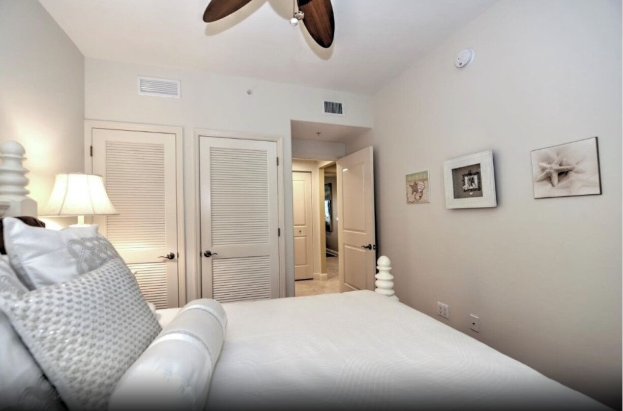 Une chambre à coucher comprenant un lit, un ventilateur de plafond, une table de chevet, une lampe et une porte donnant sur une autre pièce.