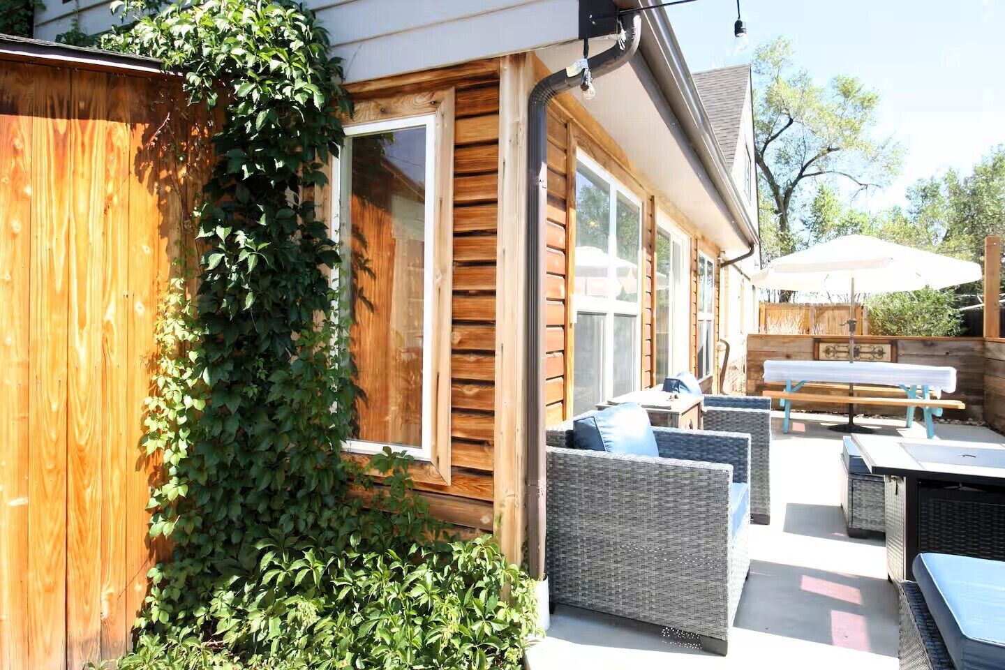 A wooden exterior wall with a window, a covered outdoor seating area with wicker furniture, and a wooden fence partially covered by ivy.
