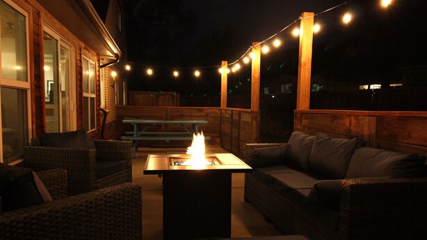Une terrasse extérieure avec des meubles en osier, une table dotée d’un foyer et des guirlandes lumineuses.