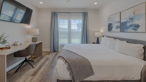 A modern hotel room with a large bed, a desk with a chair, a ceiling fan, a TV mounted on the wall, and artwork.