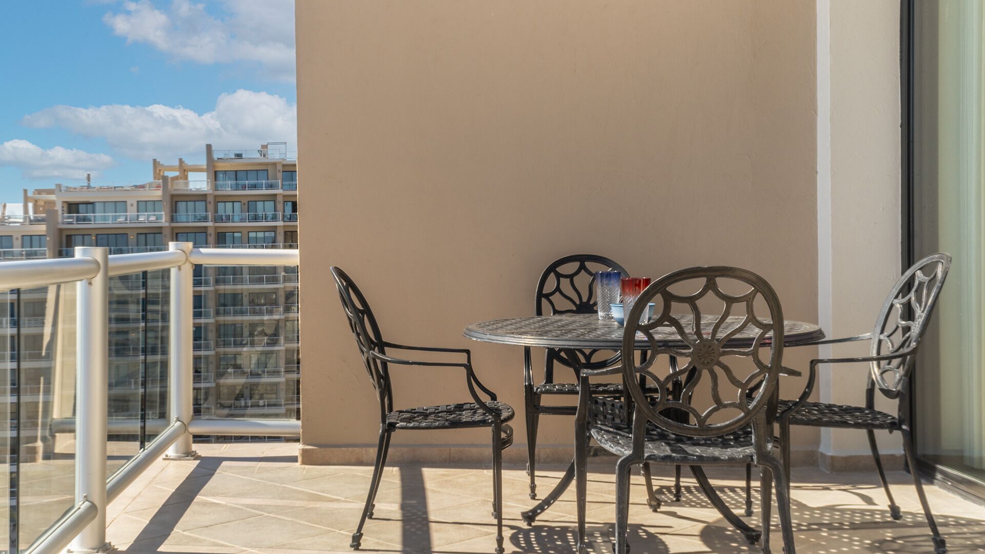 En balkon med et rundt bord og fire stole, gelænder af glas, og udsigt til høje bygninger.