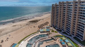 Un hotel de gran altura con vistas a la playa, una zona de piscina con tumbonas y una playa arenosa.