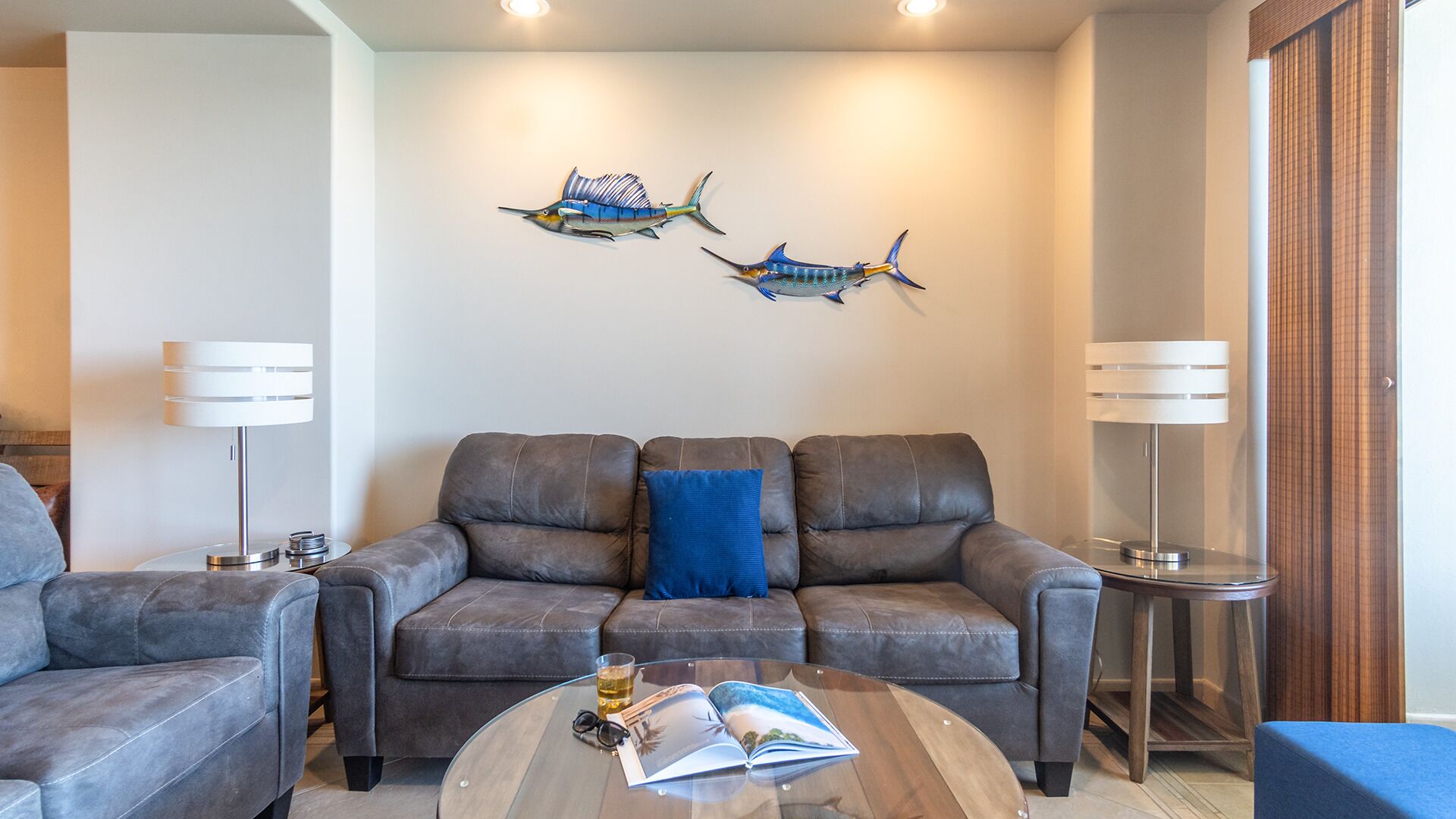 A modern living room with a grey leather sofa, a coffee table with a book and glasses, and two wall-mounted lamps.