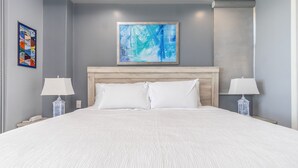 Un lit bien fait, avec du linge de lit blanc, dans une pièce aux murs gris, dotée d’une tête de lit en bois et de deux tables de chevet avec des lampes.