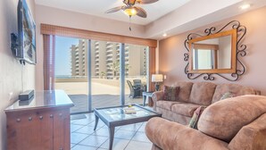 A spacious living room with a sofa, coffee table, and a large mirror. A television is mounted on the wall, and there is a balcony with a view of high-rise buildings.
