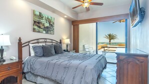 A bedroom with a bed, bedside lamps, a painting, and a view of a balcony with a palm tree.