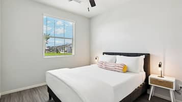 Um quarto com cama, janela com persianas, ventoinha no teto e mesa de cabeceira com abajur.