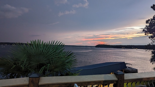 Zonsondergang vanaf een terras, met een palmpje op de voorgrond, met uitzicht op een kalm wateroppervlak en een landmassa in de verte.