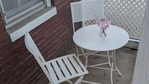 Un patio con una mesa blanca y dos sillas blancas, un jarrón pequeño con flores, y un muro de ladrillo.