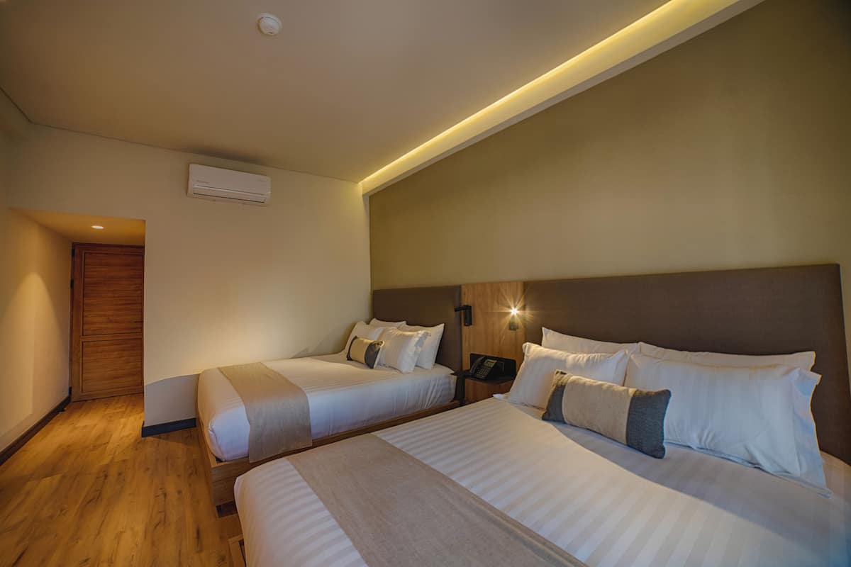 A hotel room with two beds, a wooden door, an air conditioning unit, and a wall-mounted light fixture.