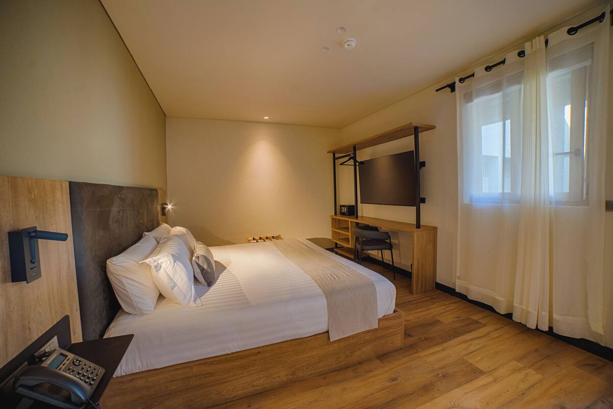 A modern hotel room with a large bed, a desk, a chair, a TV, and a wooden floor.